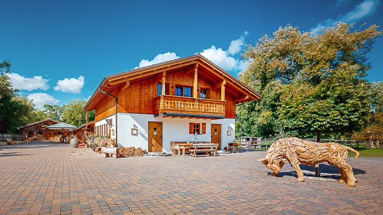 Holzhaus mit Balkon und Blumenkästen am gepflasterten Hof, davor eine Stierskulptur aus Holz